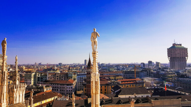 Statue atop Milan's Duomo overlooking the city's skyline, featuring modern and historical buildings under a clear blue sky.