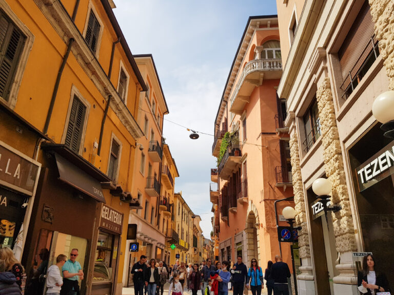 A narrow street in Verona lined with brightly colored buildings, with people walking under the warm sun and small cafes nestled between storefronts.