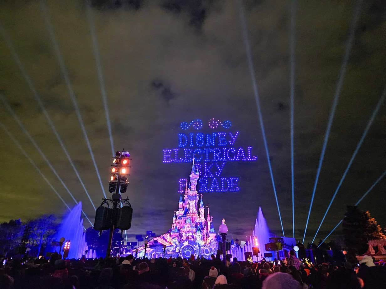 A magical nighttime spectacle at Disneyland Paris, featuring the "Disney Electrical Sky Parade" written in lights above the illuminated Sleeping Beauty Castle, with beams of light cutting through the cloudy sky.