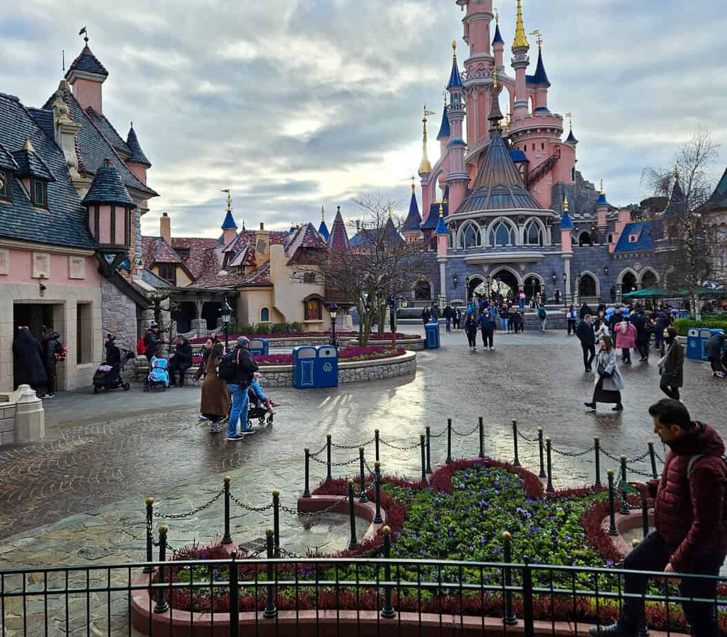 Early morning visitors stroll through the charming square in front of the Sleeping Beauty Castle at Disneyland Paris, as the cobblestone paths glisten under the soft light.
