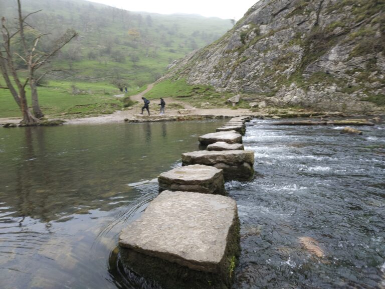 A close-up of the stepping stones leading across the clear, rippling water of a river, with grassy hills and rocky outcrops in the background.