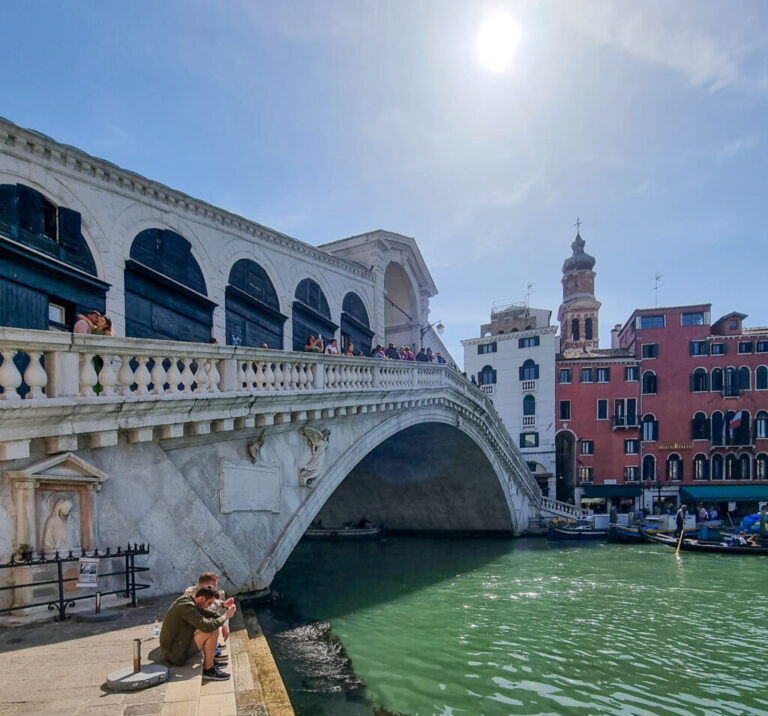Image of the Rialto Bridge in Venice, with tourists standing along the bridge and a few people seated by the green canal water below, enjoying the sunny day.