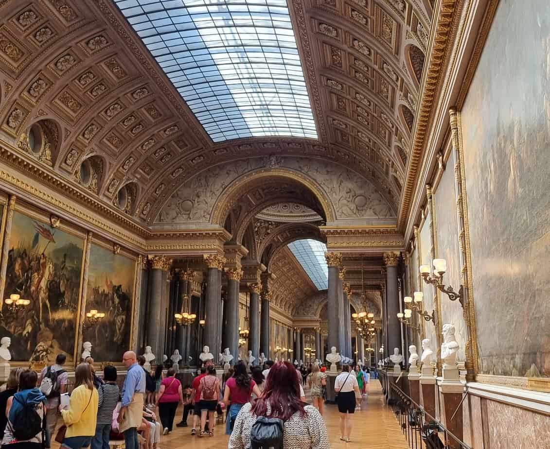 Tourists walk through an opulent hall in the Palace of Versailles, lined with grand paintings, marble busts, and a richly detailed arched ceiling with a skylight above. Exploring Versailles is a perfect addition to your 3 days in Paris itinerary.