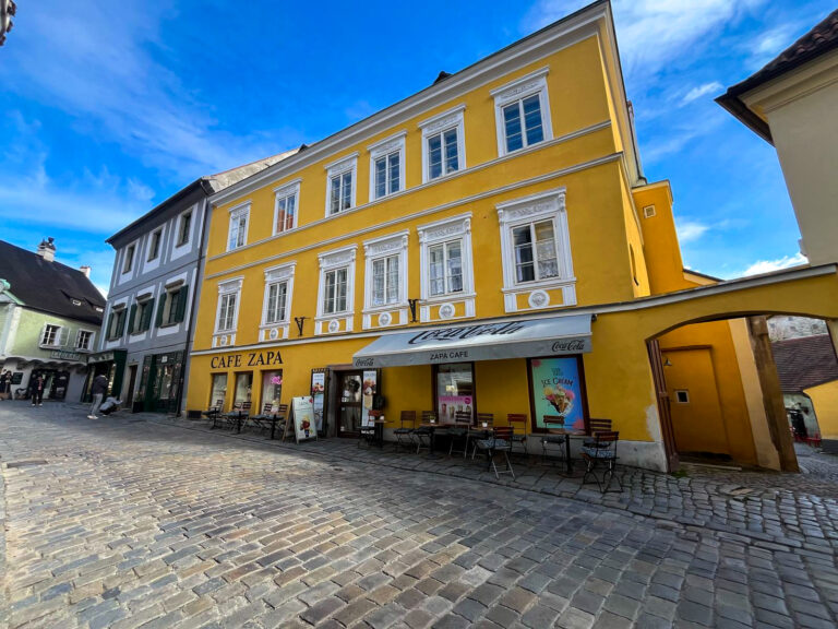 Bright yellow building housing Café Zapa in a picturesque cobblestone street, with a few chairs and signs out front, and bold blue skies overhead—an inviting stop on a relaxed Český Krumlov day trip from Prague.