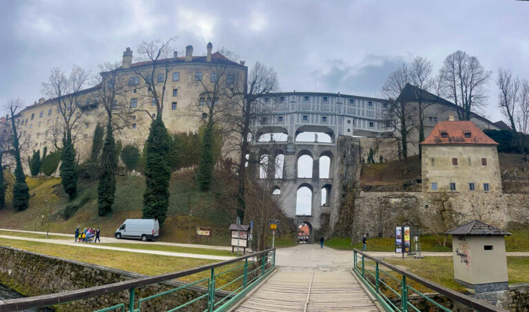 View of the Cloak Bridge and Český Krumlov Castle complex, showing its layered stone arches and towering structures nestled among bare winter trees.