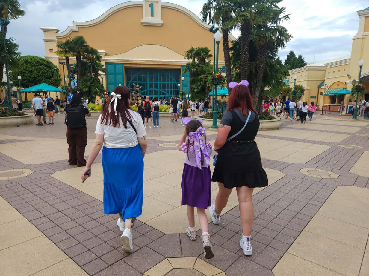 Three guests, including a child in a Rapunzel costume and two adults with Disney accessories, walk toward the entrance of Walt Disney Studios Park surrounded by palm trees and crowds—a candid view of the welcoming vibe at plus size Disneyland Paris.