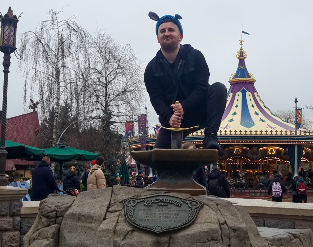 A guest poses while attempting to pull the Sword from the Stone in Fantasyland, with the carousel and castle-themed structures in the background.