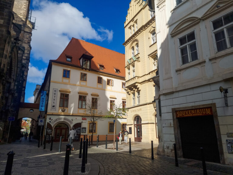 Quiet corner of Old Town Prague with historic hotels, arched walkways, and a red-roofed building basking in bright sunlight.