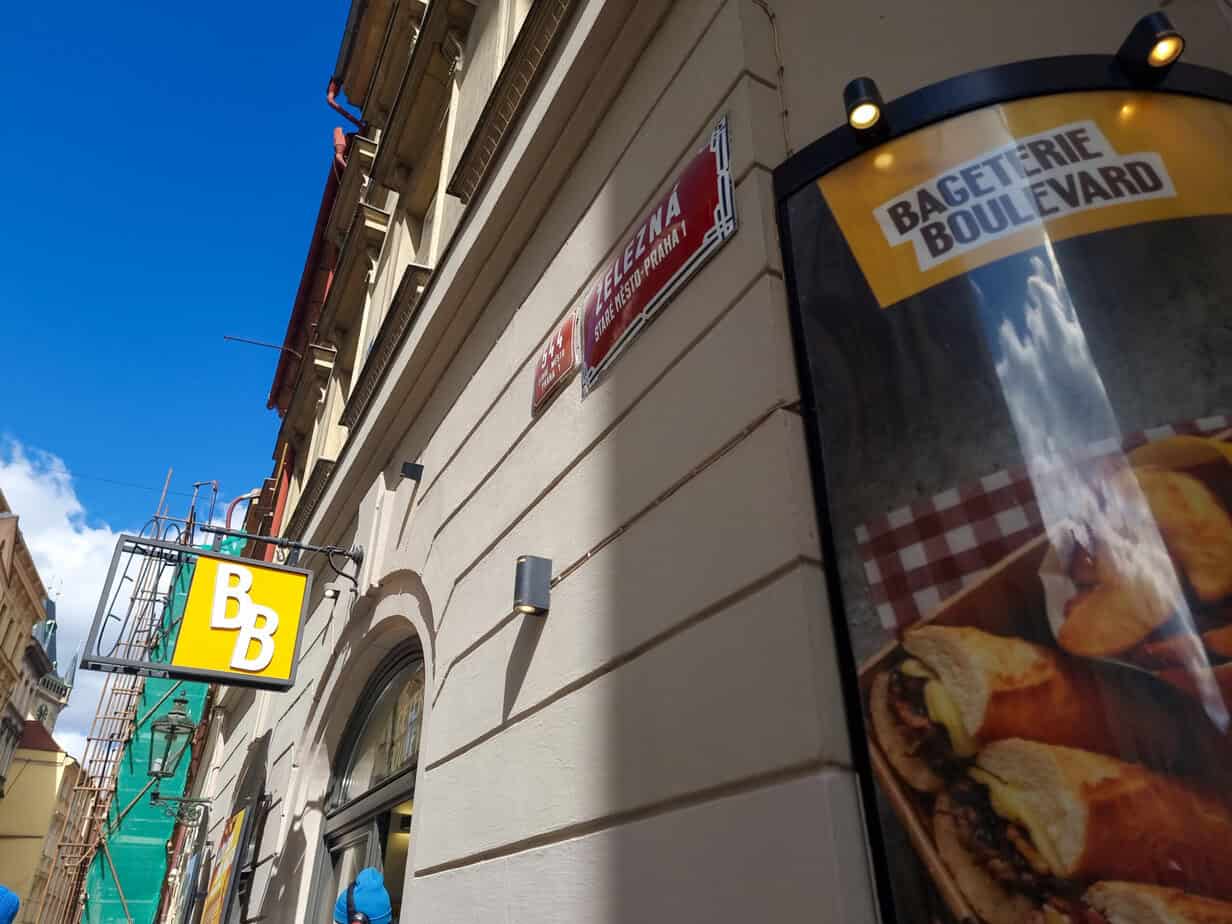 Exterior view of Baguette Boulevard café on Jindřišská Street in Prague, with a bright yellow “BB” sign and a poster featuring hot baguette sandwiches.