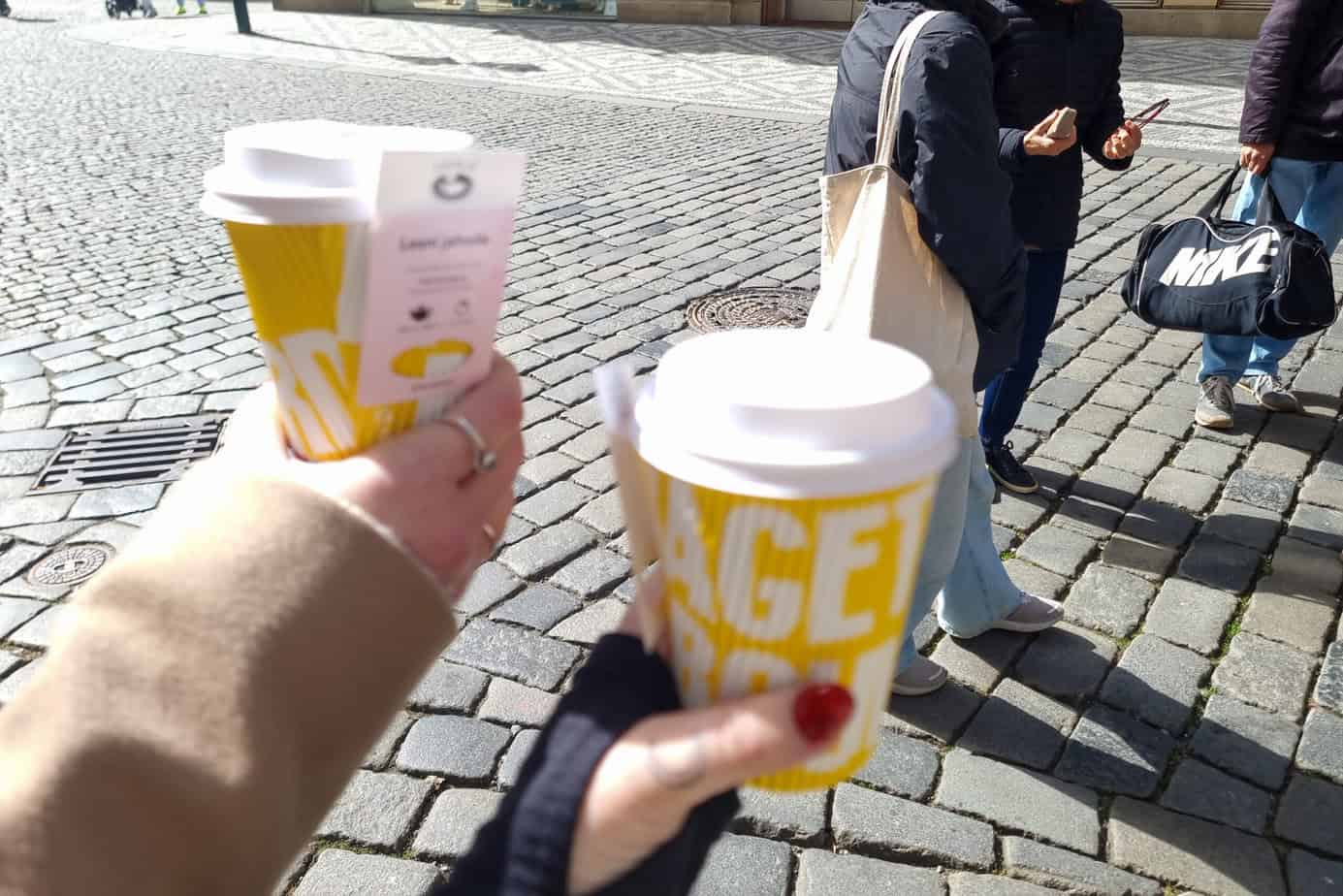 Two hands hold yellow takeaway coffee cups with sleeves that read “GET” and “YOU” while people walk across cobblestone streets in the background.