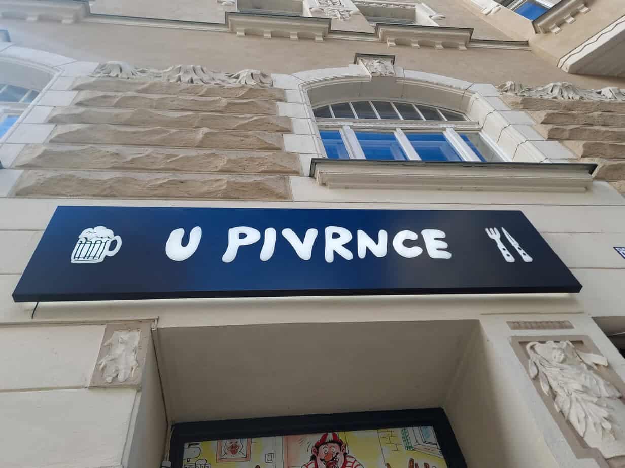 Bold navy sign for U Pivrnců pub, featuring beer and cutlery icons, mounted on a stone building in Prague's Old Town.