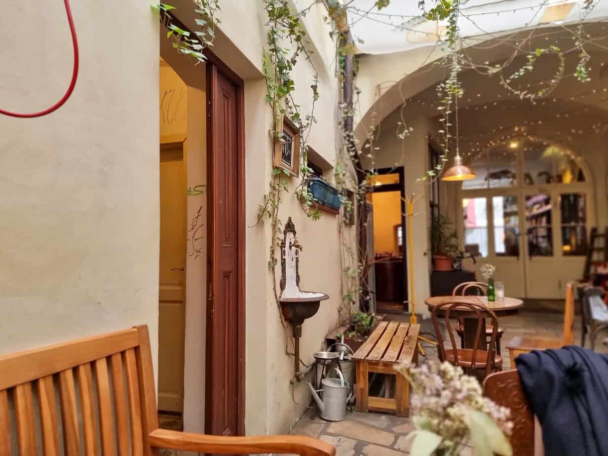 Quaint courtyard café with warm wooden seating, fairy lights overhead, a vintage wall sink, and potted plants adding to the intimate vibe—perfect for a break during 1 day in Prague.