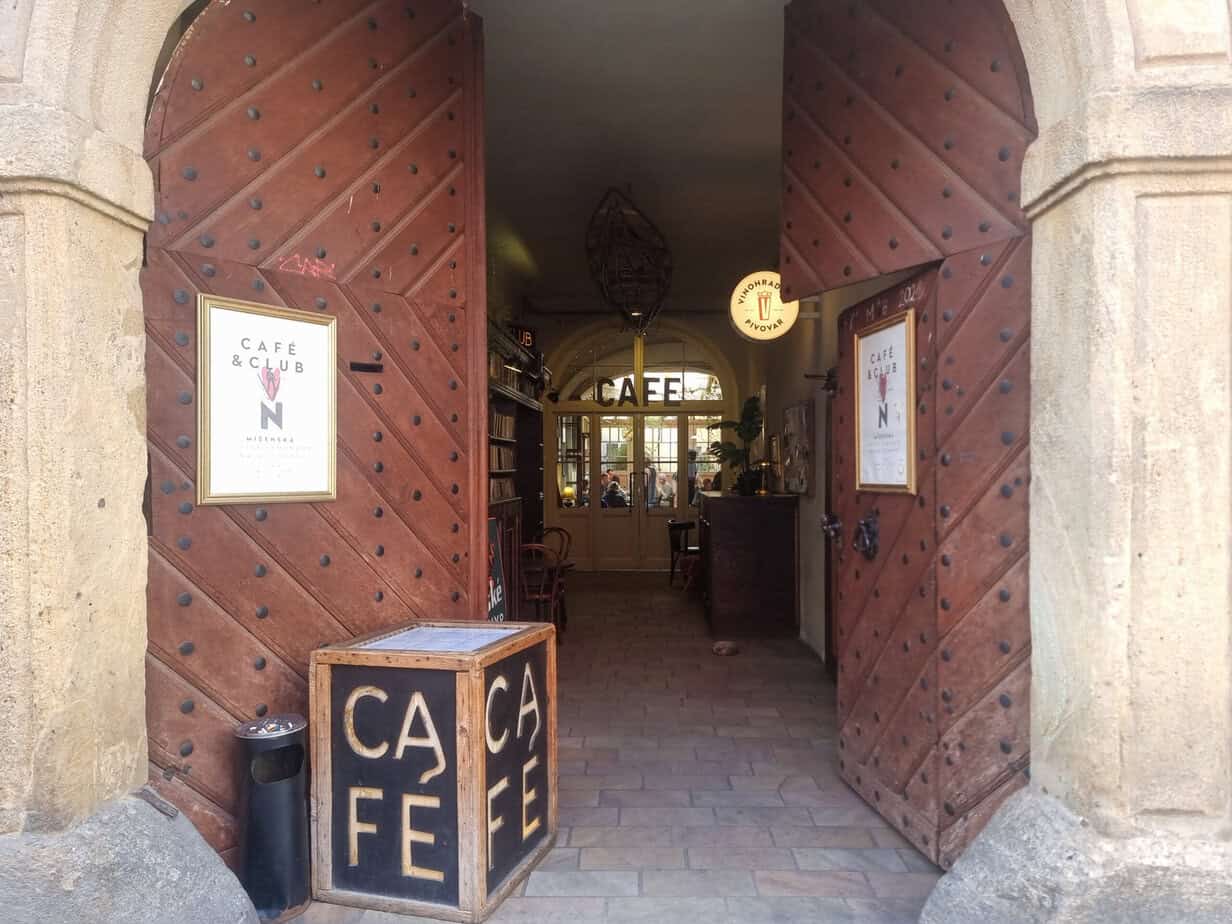 Open wooden doors lead into Café & Club N, a cozy Prague café with a rustic entrance, signboards, and bookshelves visible inside.