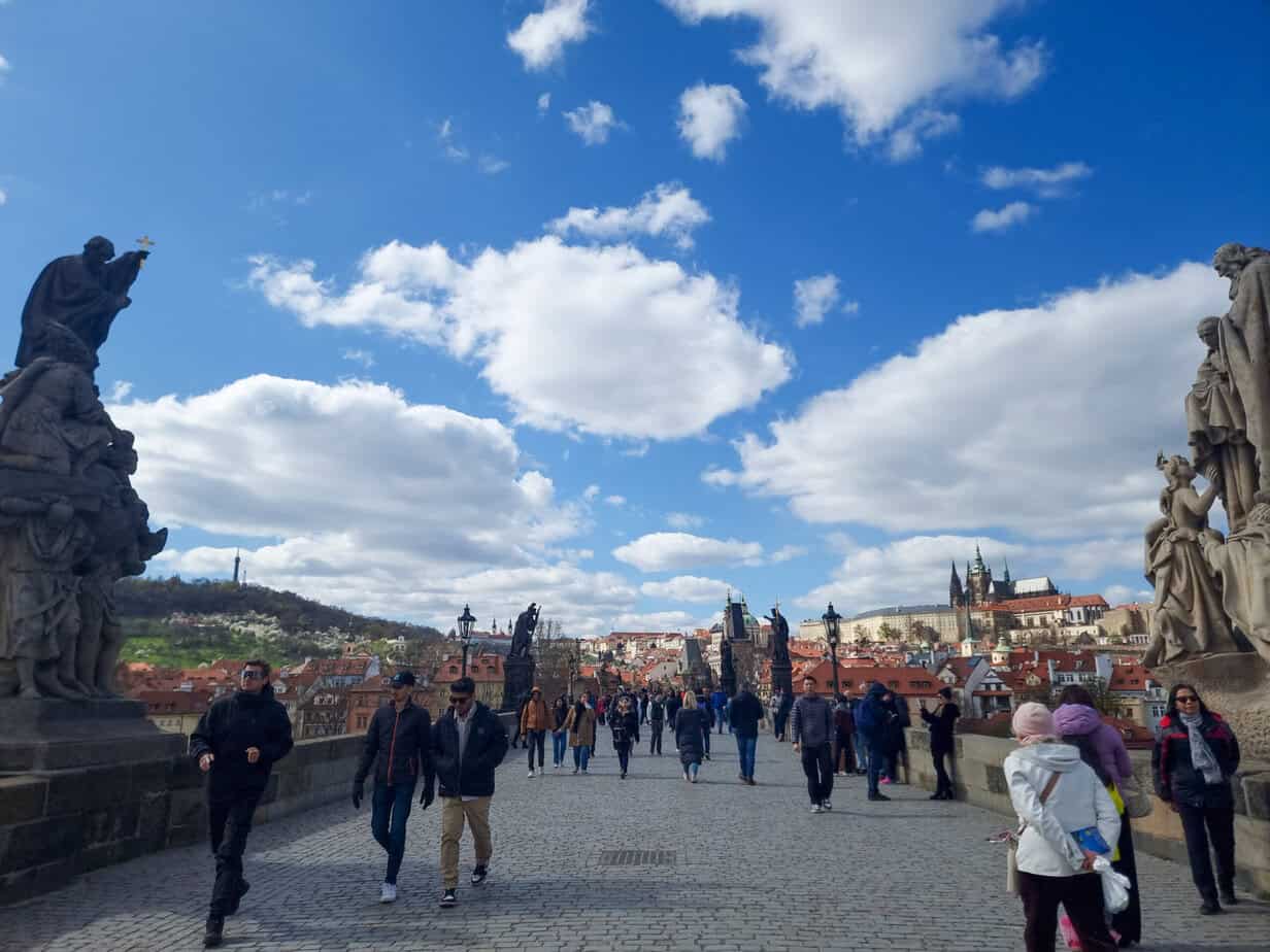 Tourists cross the Charles Bridge on a bright, breezy day with clear skies and statues towering on both sides—one of the must-see stops during 1 day in Prague.