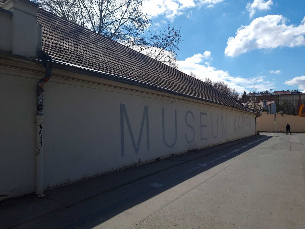 Exterior of the Museum Kampa in Prague with large gray lettering on a cream wall, captured under clear skies and surrounded by bare trees. One of many peaceful spots worth seeing in 1 day in Prague.