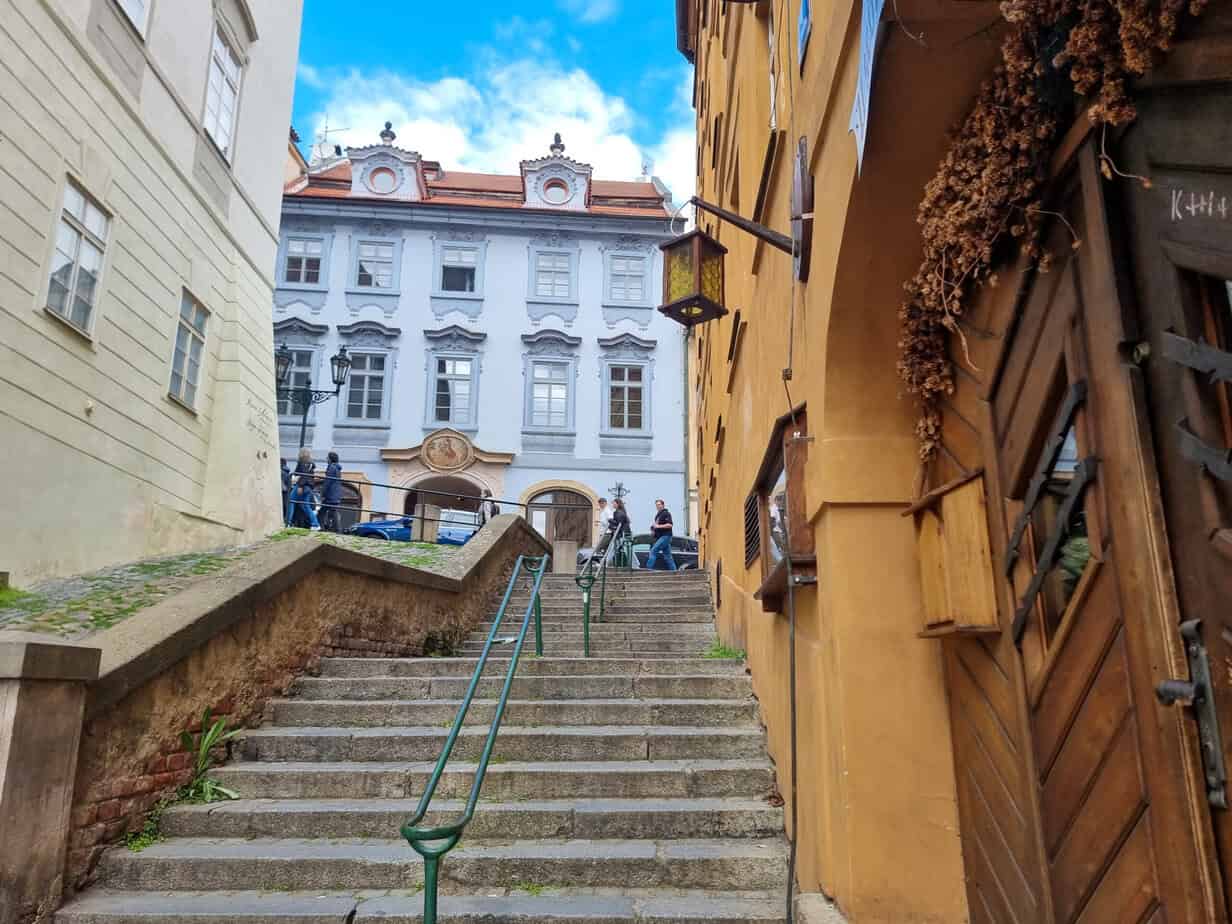 Stone stairway lined with colorful buildings leading up to a white, ornate facade in Prague. A few visitors pause at the top under a bright blue sky.