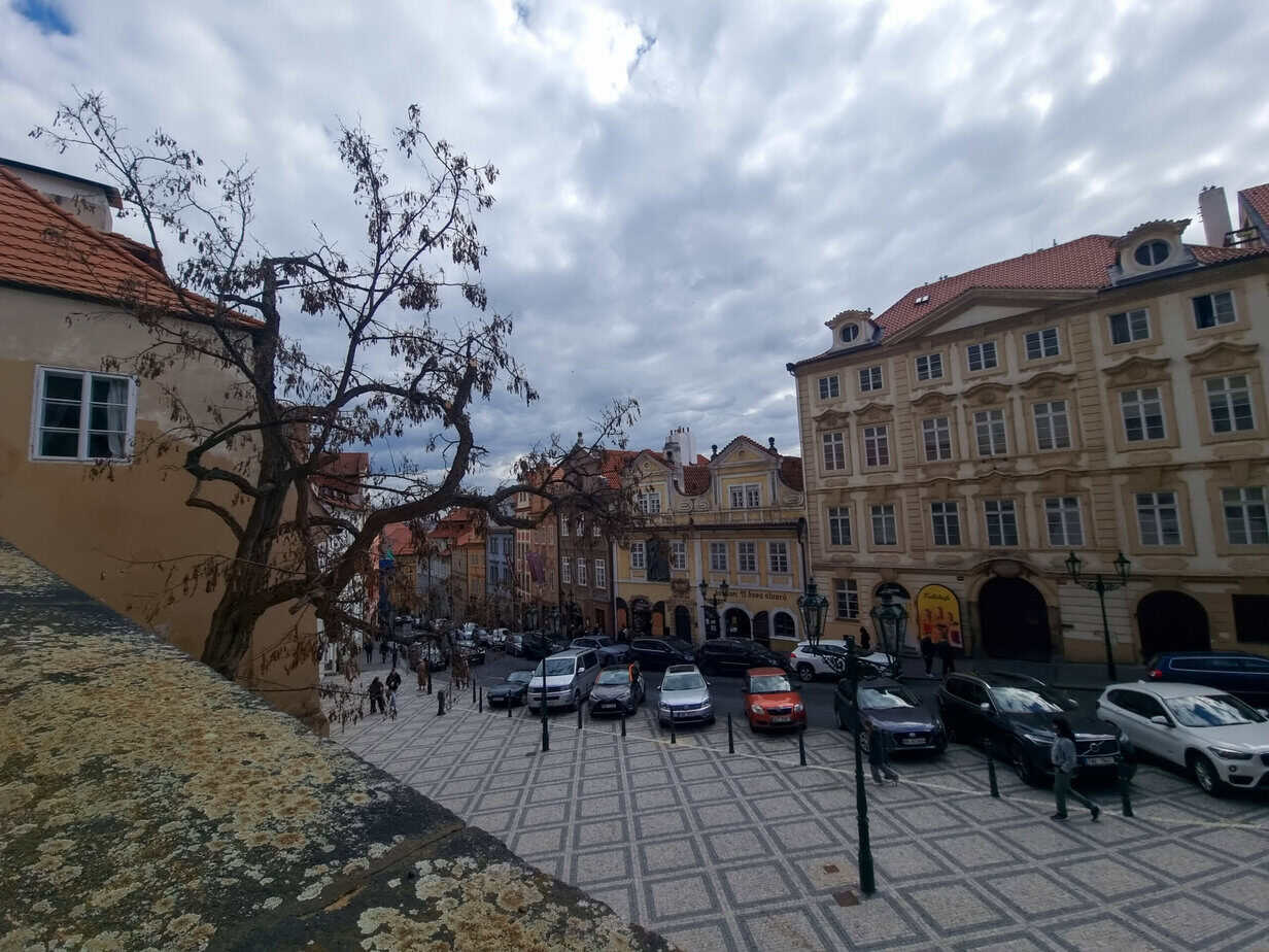 View of a quaint Prague square with baroque buildings, parked cars, and a leafless tree casting shadows over the checkered cobblestone plaza.