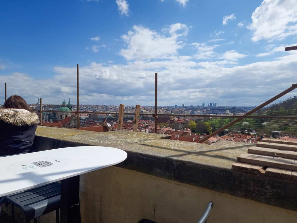A woman sits at a rooftop café table overlooking a wide panoramic view of Prague’s skyline, with red rooftops, green domes, and distant skyscrapers under a blue sky.