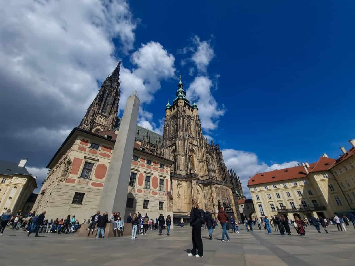 Grand view of St. Vitus Cathedral and Prague Castle against a brilliant blue sky, surrounded by lively tourists and Gothic architecture in the main castle courtyard.