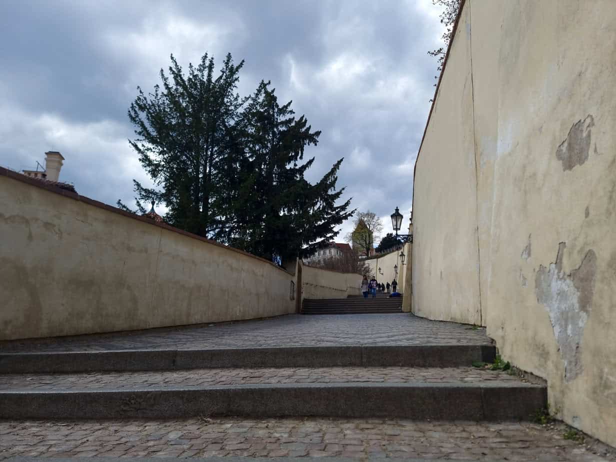Narrow cobblestone path lined with tall, weathered beige walls and a staircase leading up to a distant group of visitors. Quiet charm like this is easy to miss with only 1 day in Prague.
