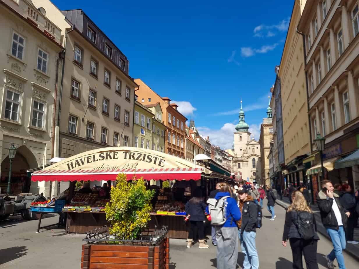 Bustling Havel Market in Prague with fruit stalls under a striped canopy, surrounded by pastel buildings and topped with the sign “Havelské Tržiště.”