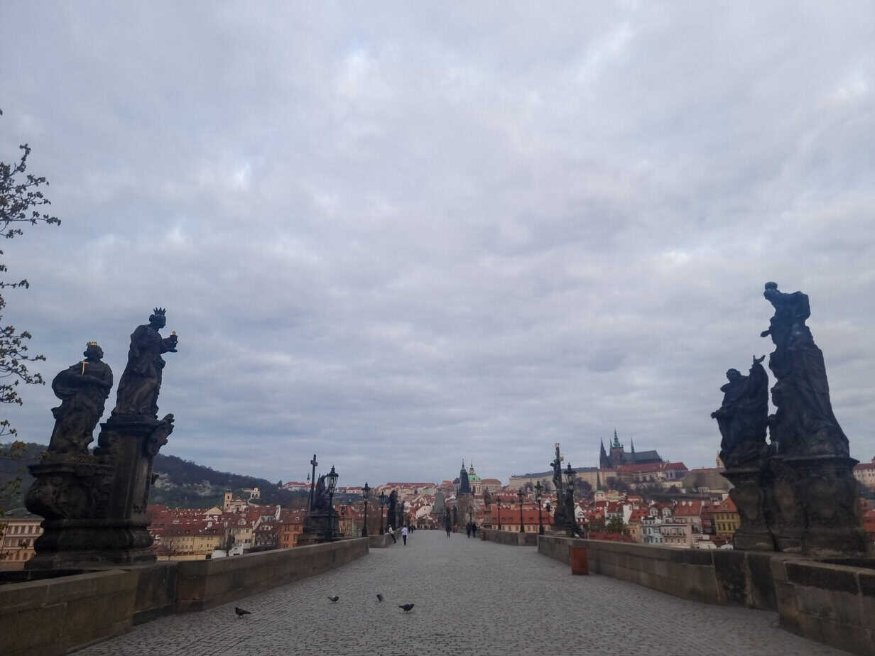 View down the iconic Charles Bridge lined with statues, looking toward Prague Castle and the red-roofed skyline under a soft overcast sky—an essential experience during 1 day in Prague.