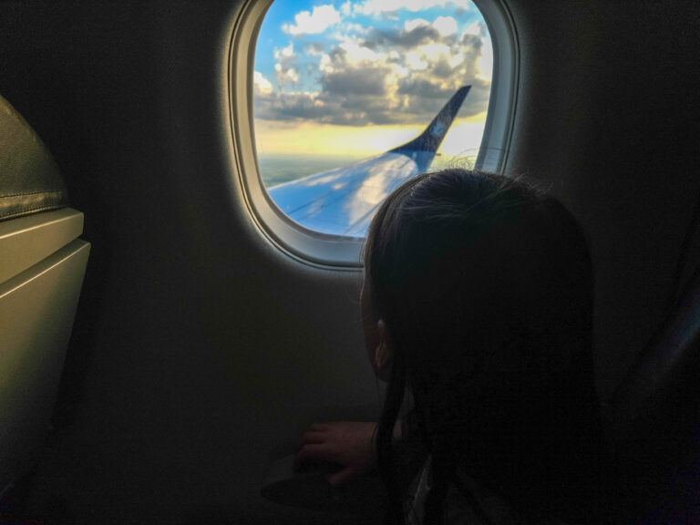 A child gazing out of an airplane window at sunset, watching the wing and sky during a flight towards adventure.