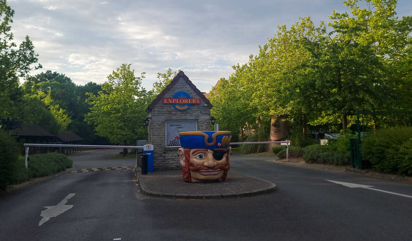 A quaint entrance to the Explorers Hotel with a whimsical pirate head sculpture at the gate and a sign above reading “Explorers.” Trees and a small booth complete the charming and playful atmosphere.