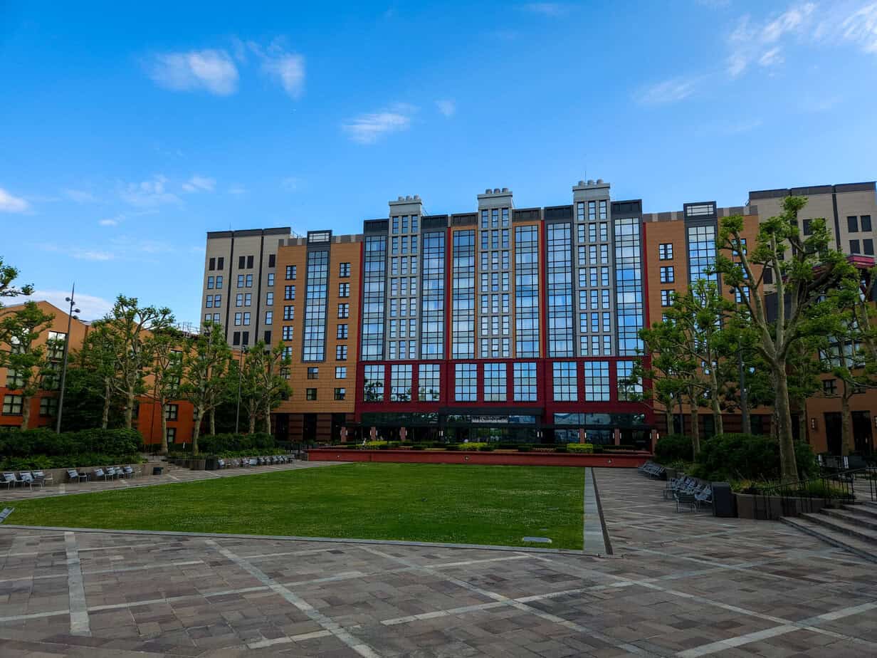 Exterior of Disney Hotel New York – The Art of Marvel, showcasing modern architecture with red and beige tones, large reflective windows, and a central grassy courtyard.