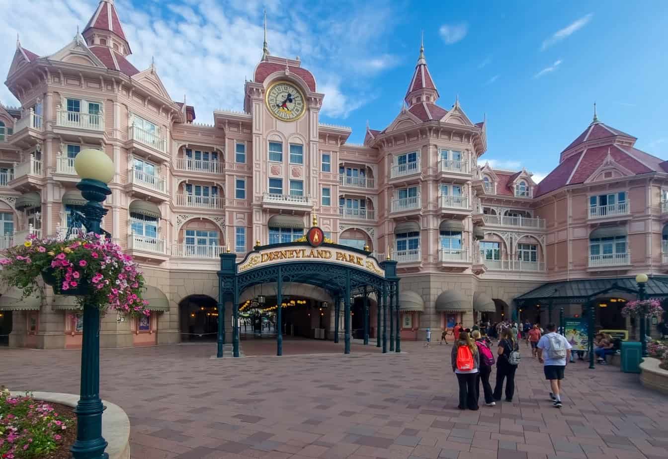 The entrance to Disneyland Park in Paris with the iconic pink Victorian-style Disneyland Hotel in the background and guests walking toward the gates beneath a golden sign.
