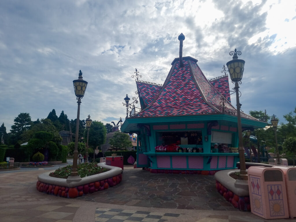 A brightly colored, fairy-tale-themed snack stand with a red-and-pink shingled roof and teal walls, surrounded by lantern-style lights and floral planters.