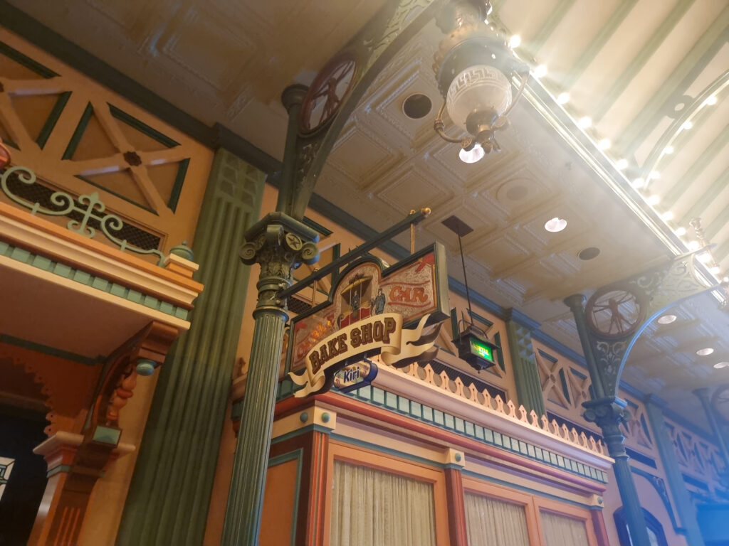 Inside a Victorian-style arcade, a decorative overhead sign for Victoria’s “Home-Style” Bake Shop hangs beneath an old-fashioned ceiling.
