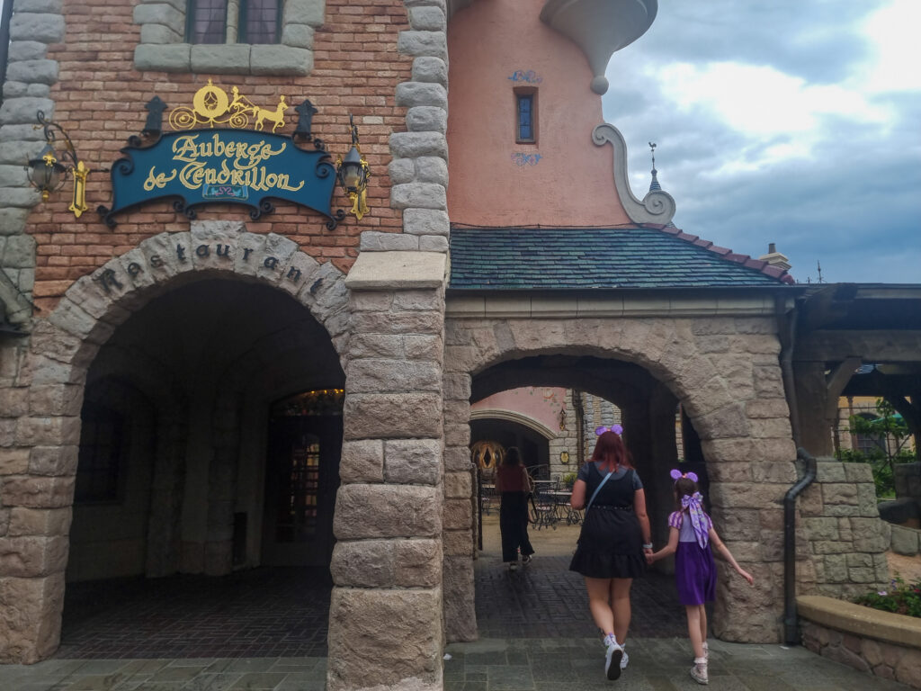 A stone archway and turreted entrance to Auberge de Cendrillon, a Cinderella-themed dining experience, as two guests walk through, both wearing Disney-themed outfits.