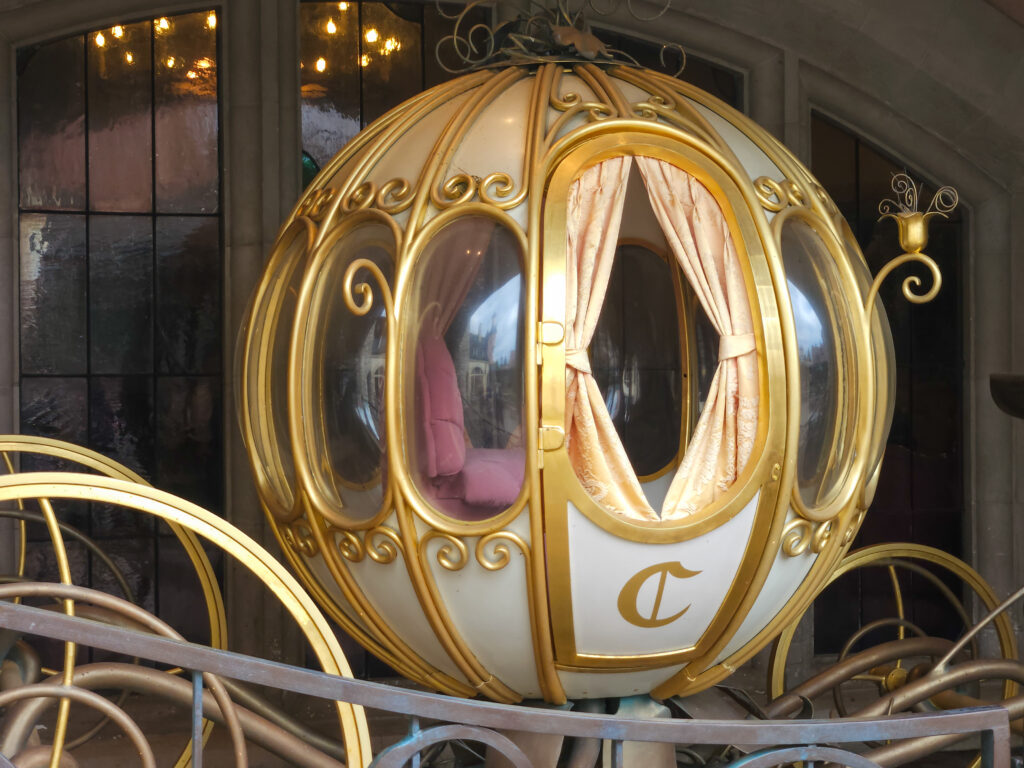 A decorative white and gold pumpkin-shaped carriage with glass panels and the letter "C" on the door, evoking Cinderella’s magical ride.