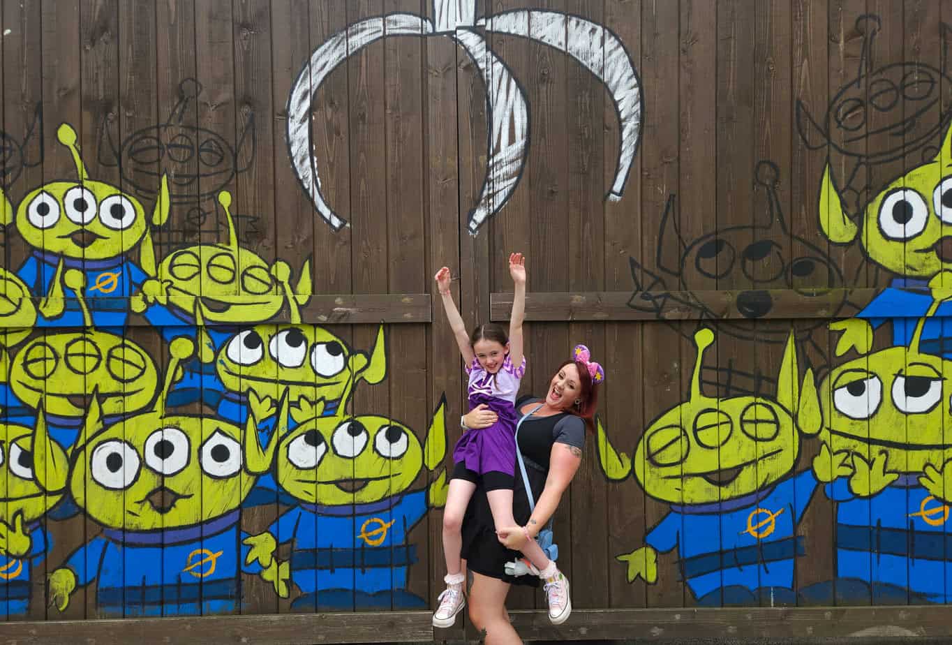 A woman lifts a child with raised arms beneath a painted claw machine mural of green Toy Story aliens — fun and playful street art within the park.