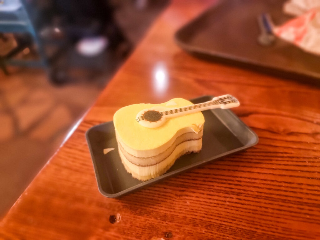 A close-up of a dessert shaped like an acoustic guitar, served on a tray atop a wooden table inside a Disneyland Paris dining area.