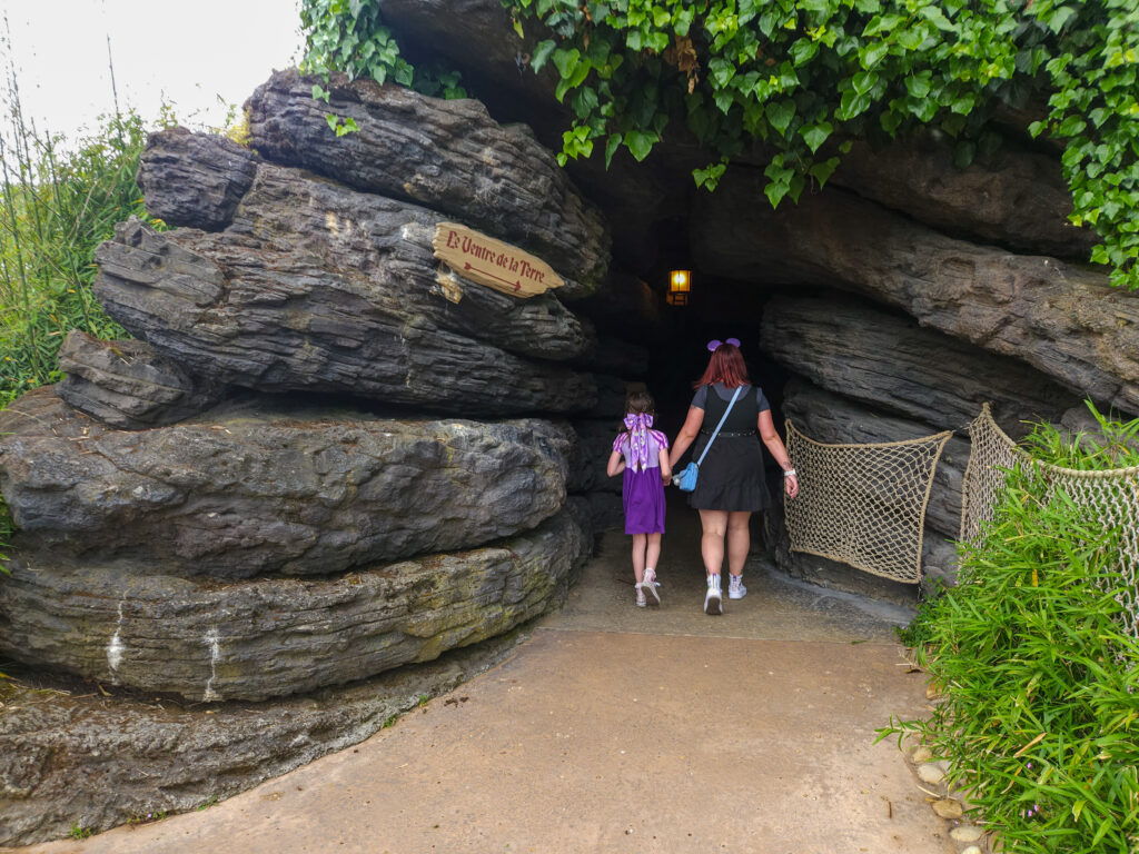 A woman and a young girl enter a rocky cave tunnel at Adventure Isle, surrounded by greenery and marked with a wooden sign.