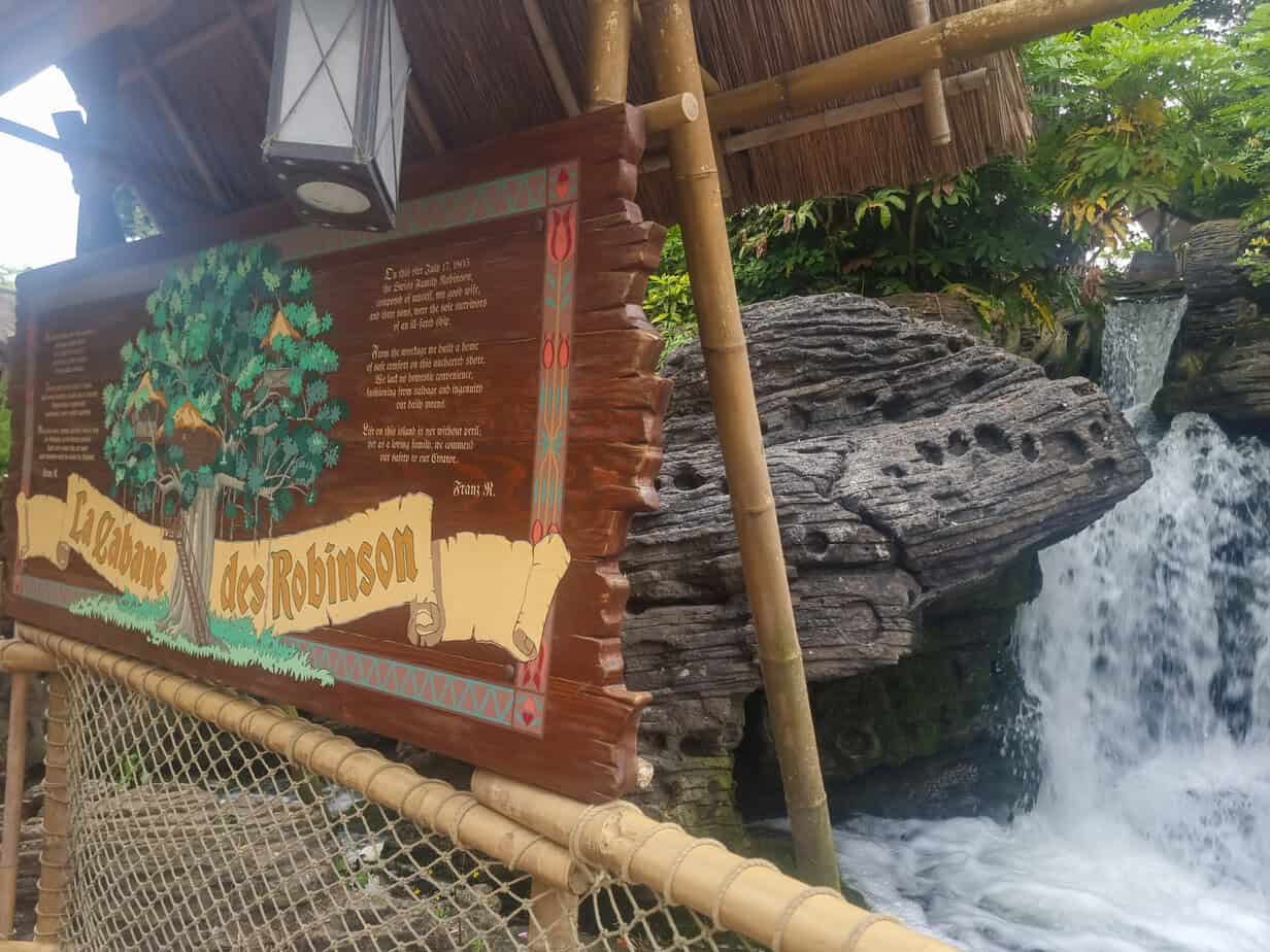 A colorful wooden sign for "La Cabane des Robinson" stands beside a rushing waterfall framed by tropical plants — a peaceful corner among disneyland paris secrets.