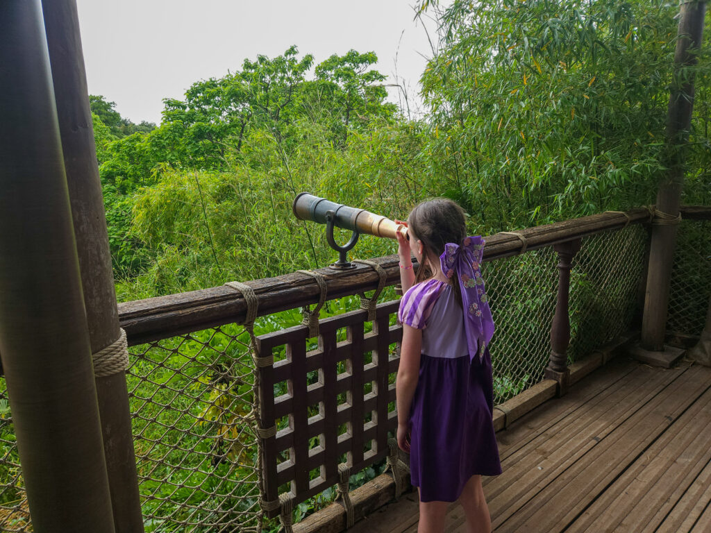 A young girl in a purple dress peers through a mounted telescope from a wooden lookout platform nestled in lush greenery.