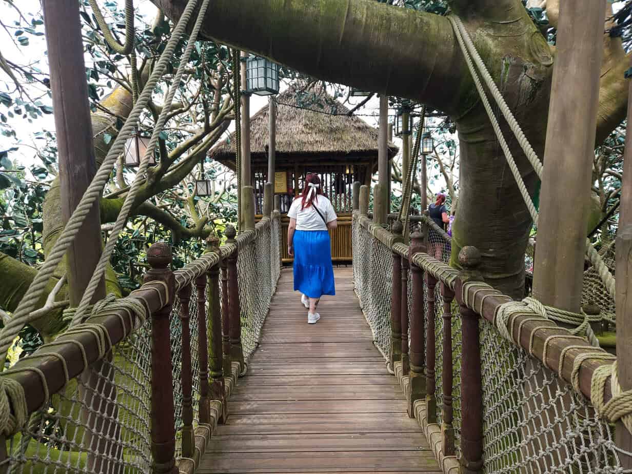 Guests walk along a wooden rope bridge surrounded by enormous tree branches, heading toward a thatched-roof lookout in Adventureland.