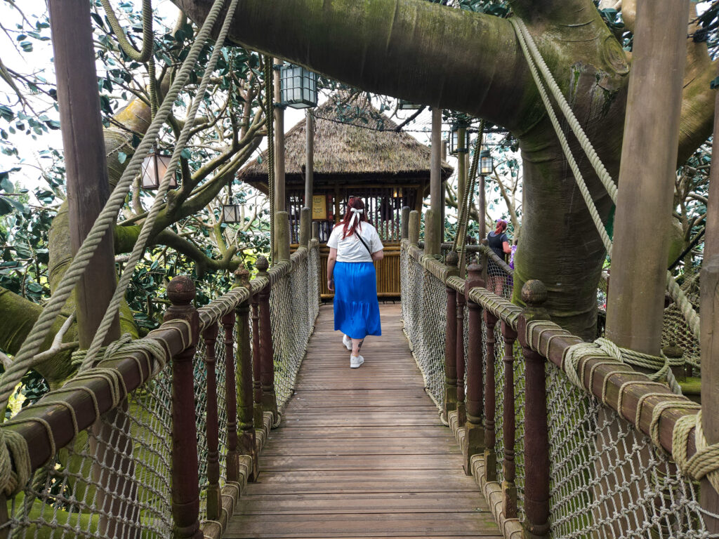 A guest walks across a wooden suspension bridge wrapped in ropes and nestled among giant tree trunks in Adventure Isle.