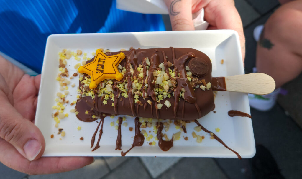 Close-up of a chocolate-dipped Magnum ice cream bar topped with chopped nuts, chocolate drizzle, and a yellow candy sheriff badge decoration, served on a white rectangular plate. This fun treat is part of the themed Disneyland Paris snacks collection.
