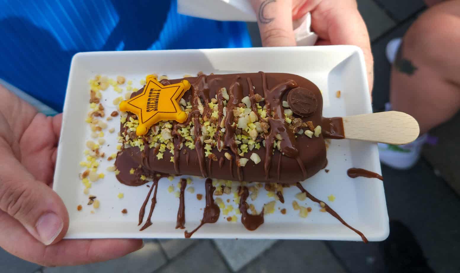 Close-up of a chocolate-dipped Magnum ice cream bar topped with chopped nuts, chocolate drizzle, and a yellow candy sheriff badge decoration, served on a white rectangular plate. This fun treat is part of the themed Disneyland Paris snacks collection.