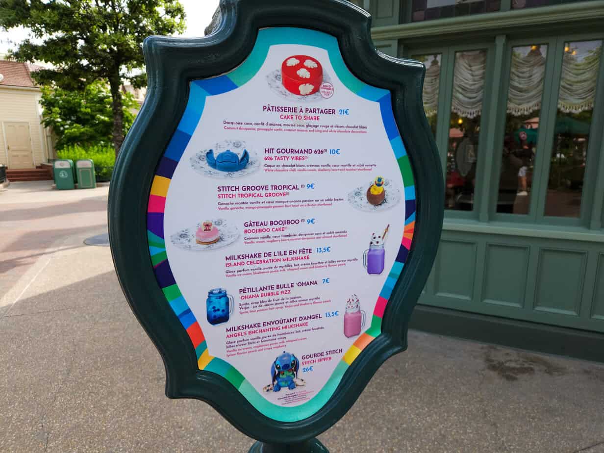 A colorful snack menu board outside a Disneyland Paris eatery displaying treats like Stitch Groove Tropical, Boojiboo cake, and themed milkshakes with character-inspired decorations.