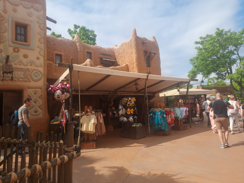 Outdoor souvenir stand in Adventureland sells Disney-themed apparel and plushies under a canopy, with tan, adobe-style architecture and visitors browsing.