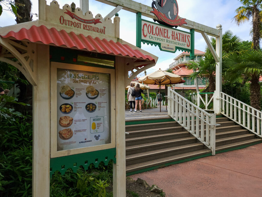 Entrance to Colonel Hathi’s Pizza Outpost with stairs leading up to a shaded patio; a posted menu shows pizza, pasta, and kid-friendly meal options.