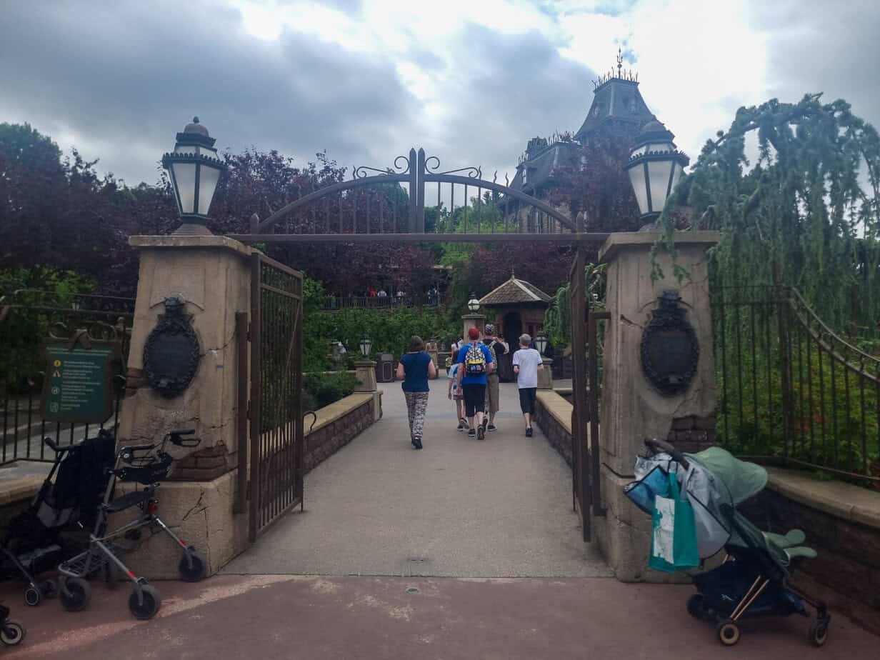Families walk through the aged, eerie gates of Phantom Manor, with overgrown foliage and empty strollers nearby adding to the mysterious vibe.