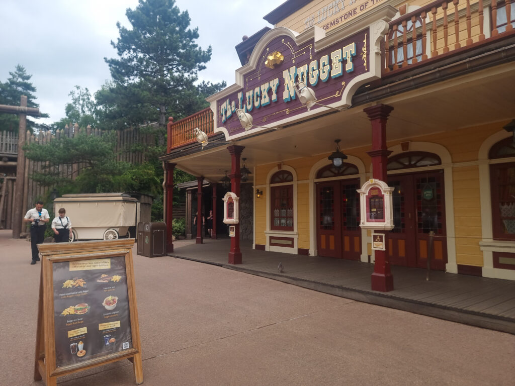 The façade of The Lucky Nugget Saloon showcases a classic Western theme with red columns, a balcony, and menu boards outside.