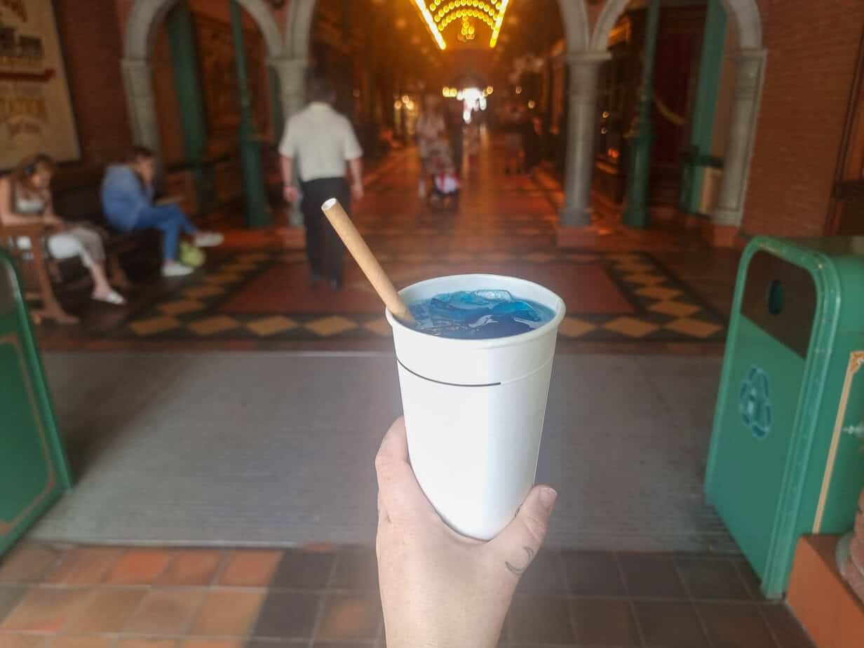 A blue slushy-style drink with a wooden straw held in front of the illuminated covered walkway inside Disneyland Paris.