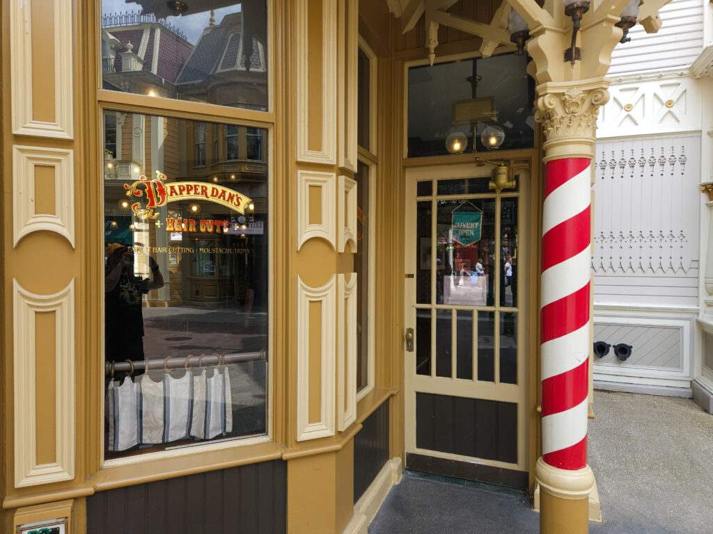 A nostalgic barbershop corner on Main Street U.S.A., with a red-and-white striped barber pole and gold lettering on the window reading “Dapper Dan’s Hair Cuts.”